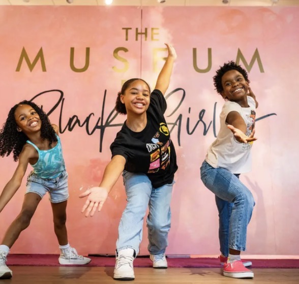 Three young Black girls dancing in front of the Museum for Black Girls signature pink wall